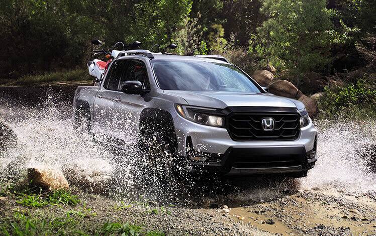 Front passenger-side view of the 2023 Honda Ridgeline Sport in Lunar Silver Metallic with available HPD™ Bronze Package shown driving through a small stream.